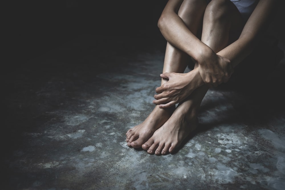 Person sitting on the floor in a dimly lit room, arms wrapped around legs, symbolizing trauma and isolation after sex trafficking.